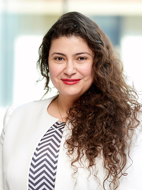 Person with long curly hair wearing a white blazer and a patterned top, standing in a bright indoor setting.